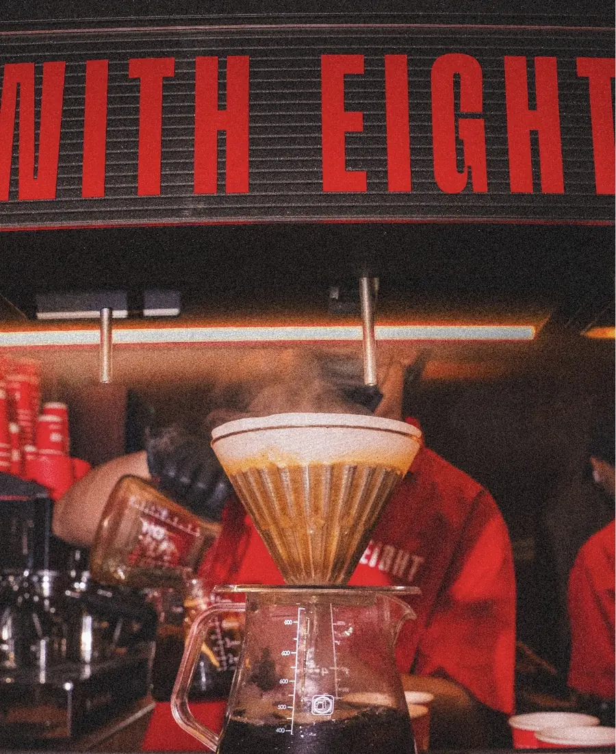 مصفاة V60 تقطّر على البار وفوقها لوحة حمراء مكتوب عليها WITH EIGHT